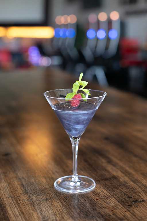 "Onyx Tini" cocktail at Jive Lounge, a striking purple-hued drink served in a martini glass, garnished with a fresh raspberry and mint sprig on a wooden table.