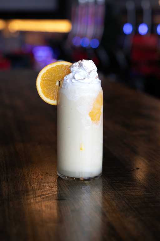"The Placebo" cocktail at Jive Lounge, a creamy citrus-infused drink topped with whipped cream and garnished with a fresh orange slice, served in a tall glass.