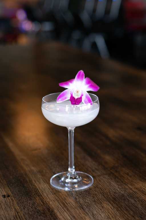"Like A Pearl" cocktail at Jive Lounge, a refreshing drink elegantly garnished with a vibrant orchid, served in a coupe glass on a wooden table.