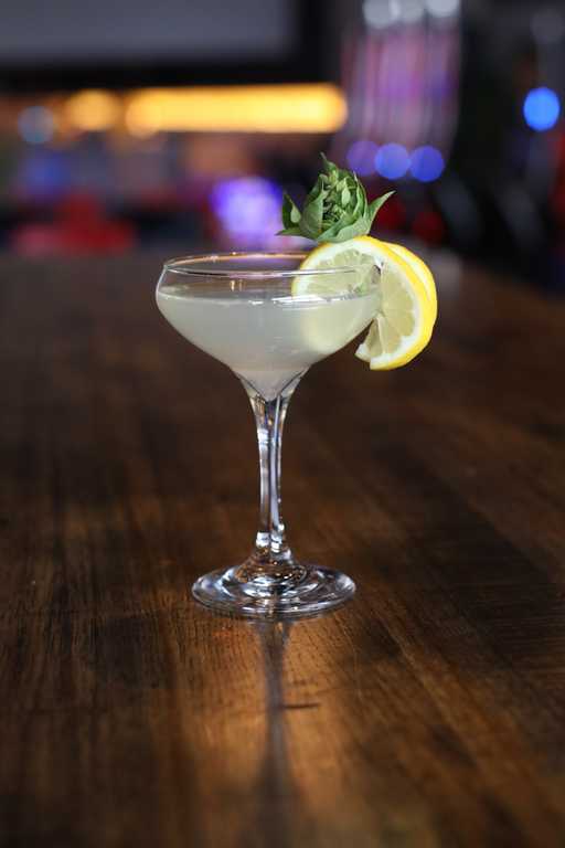 "Smoke n Spirits" cocktail at Jive Lounge, a sophisticated drink served in a coupe glass, garnished with a fresh lemon slice and basil sprig on a wooden table.